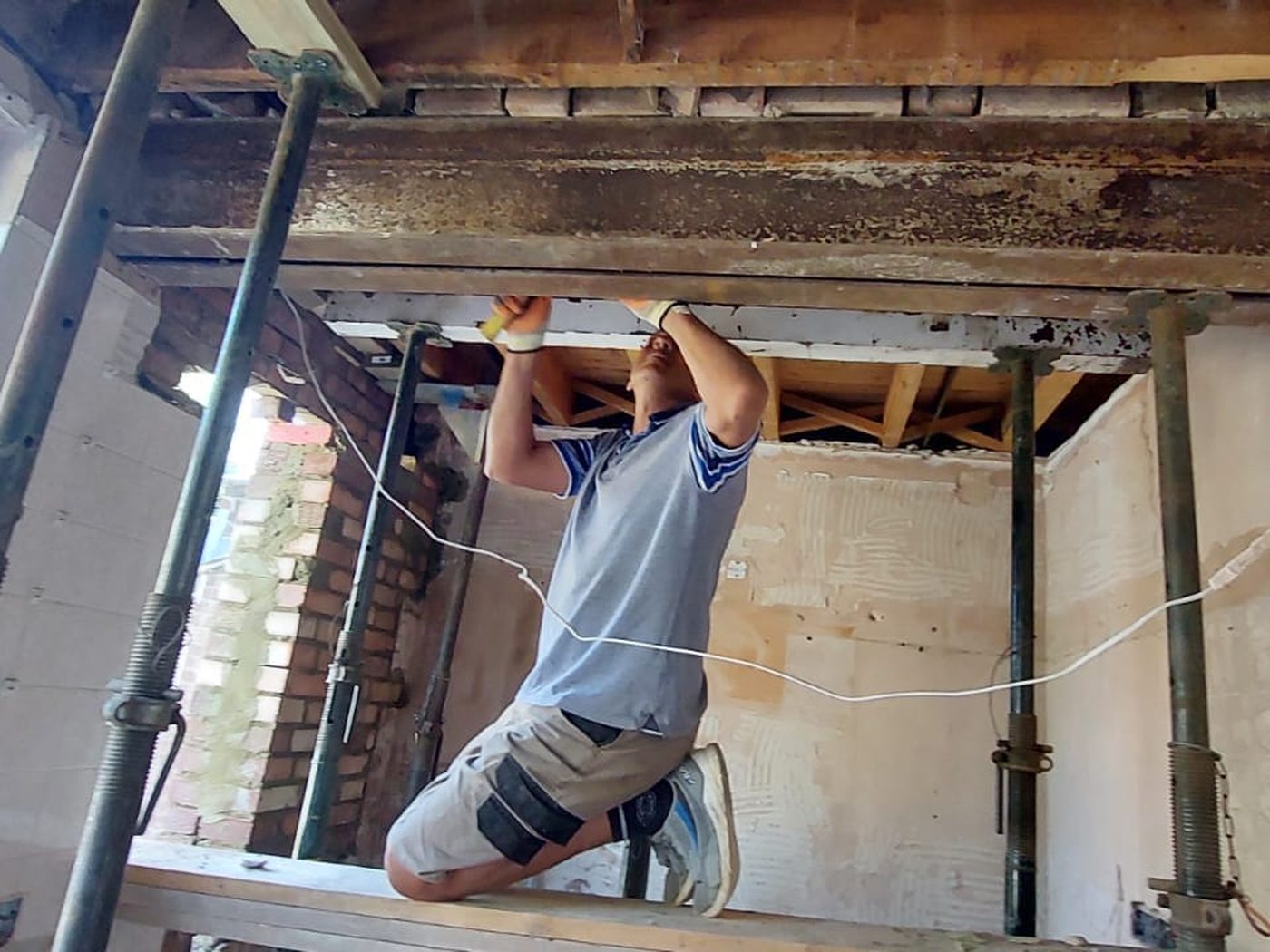 A GS Renovation tradesman working beneath an installed steel beam, with acrow props still carrying the floor above